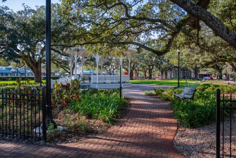 A brick path lined with mature oak trees leading to a white gazebo in League City
